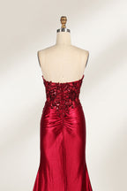 This photograph features a mannequin dressed in a strapless, floor-length, deep red satin evening gown. The gown is adorned with intricate sequin embroidery on the bodice and a high slit on the left side, revealing black stockings. The fabric has a smooth, luxurious texture. The mannequin stands on a black stool against a plain white background, creating a stark contrast that highlights the gown's vibrant color and detailed design. The overall style is elegant and glamorous, suitable for formal occasions.7
