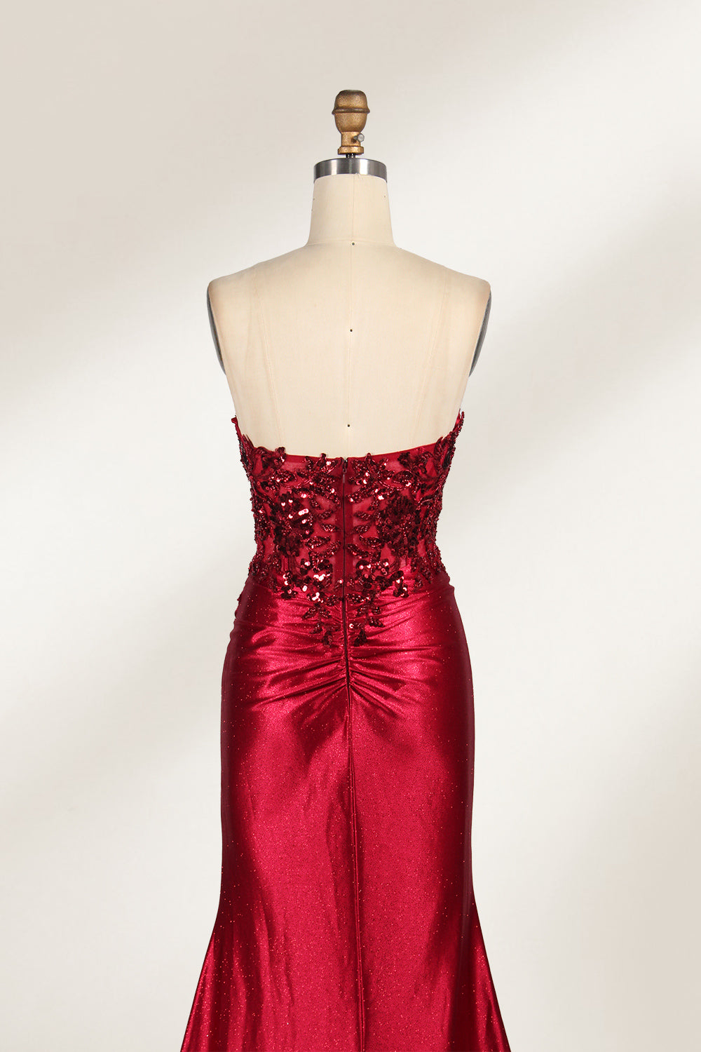 This photograph features a mannequin dressed in a strapless, floor-length, deep red satin evening gown. The gown is adorned with intricate sequin embroidery on the bodice and a high slit on the left side, revealing black stockings. The fabric has a smooth, luxurious texture. The mannequin stands on a black stool against a plain white background, creating a stark contrast that highlights the gown's vibrant color and detailed design. The overall style is elegant and glamorous, suitable for formal occasions.7