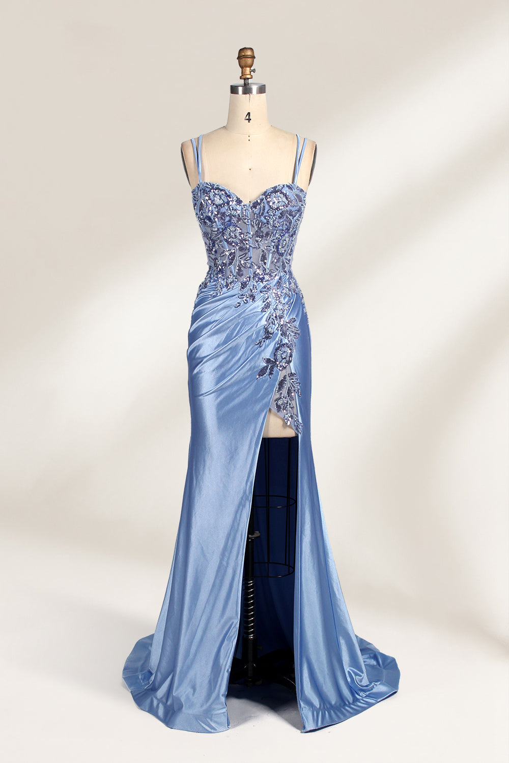 This photograph features a mannequin dressed in a luxurious, floor-length, light blue satin evening gown. The gown is intricately designed with delicate floral embroidery in silver and blue tones, primarily concentrated on the bodice and left hip. It has a plunging neckline and spaghetti straps, with a daring thigh-high slit on the left side. The mannequin is positioned against a plain, off-white background, allowing the gown's opulent texture and detailed craftsmanship to stand out. The gown exudes eleganc