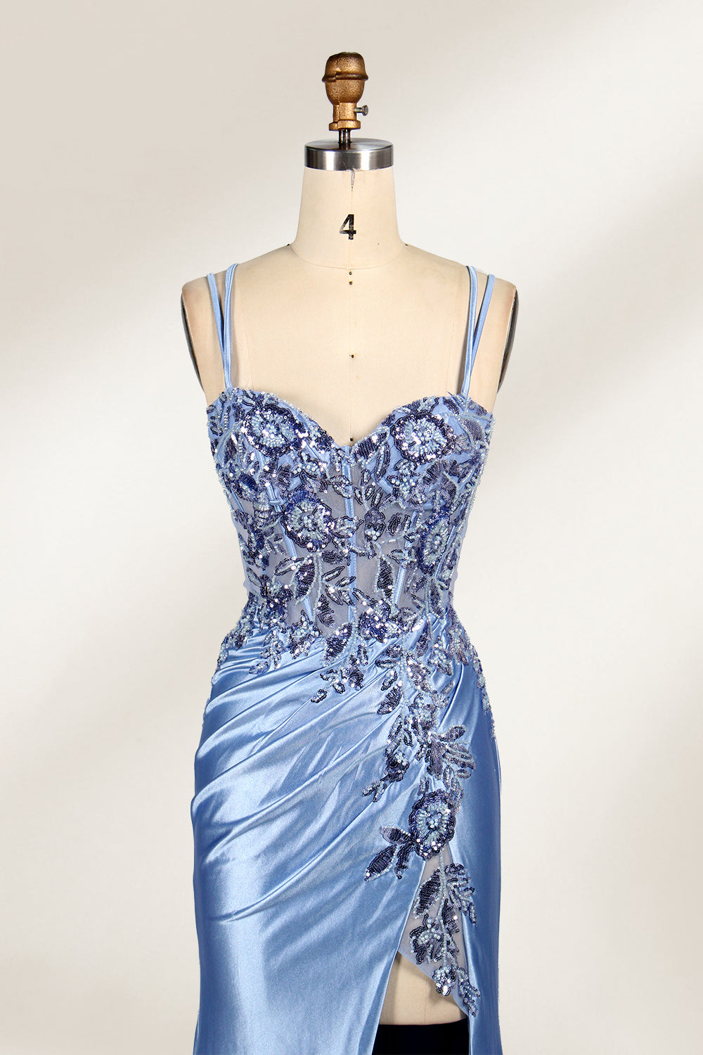 This photograph features a mannequin dressed in a luxurious, floor-length, light blue satin evening gown. The gown is intricately designed with delicate floral embroidery in silver and blue tones, primarily concentrated on the bodice and left hip. It has a plunging neckline and spaghetti straps, with a daring thigh-high slit on the left side. The mannequin is positioned against a plain, off-white background, allowing the gown's opulent texture and detailed craftsmanship to stand out. 6