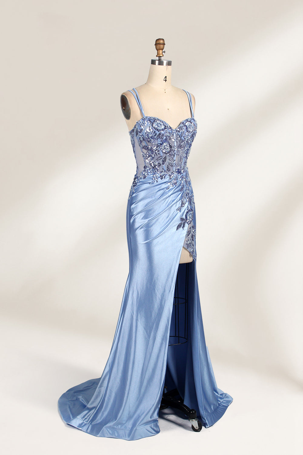 This photograph features a mannequin dressed in a luxurious, floor-length, light blue satin evening gown. The gown is intricately designed with delicate floral embroidery in silver and blue tones, primarily concentrated on the bodice and left hip. It has a plunging neckline and spaghetti straps, with a daring thigh-high slit on the left side. The mannequin is positioned against a plain, off-white background, allowing the gown's opulent texture and detailed craftsmanship to stand out. The gown exudes eleganc