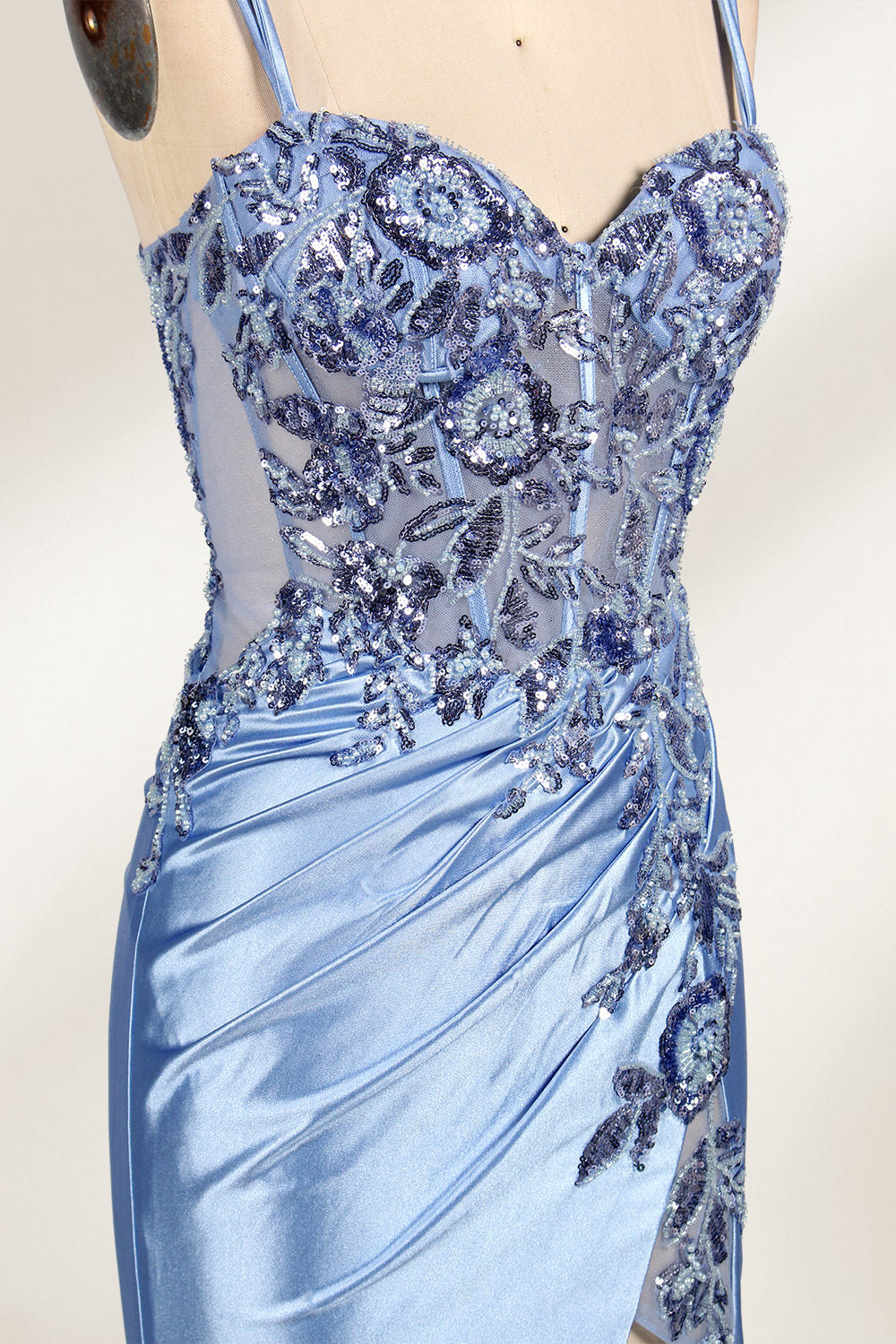 This photograph features a mannequin dressed in a luxurious, floor-length, light blue satin evening gown. The gown is intricately designed with delicate floral embroidery in silver and blue tones, primarily concentrated on the bodice and left hip. It has a plunging neckline and spaghetti straps, with a daring thigh-high slit on the left side. The mannequin is positioned against a plain, off-white background, allowing the gown's opulent texture and detailed craftsmanship to stand out. 4