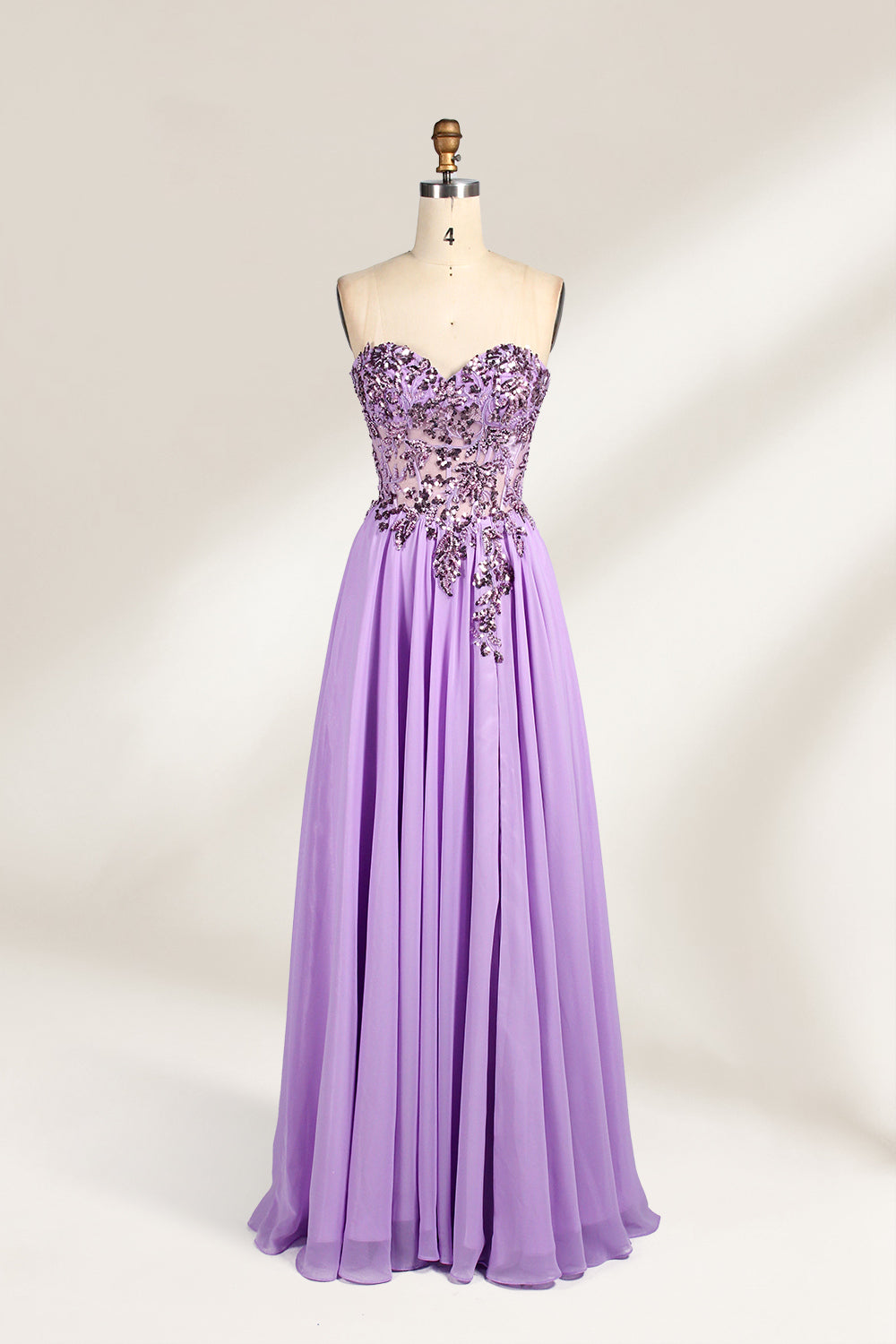 A photograph of a lavender, strapless, floor-length evening gown displayed on a dress form. The gown features intricate floral lace detailing on the bodice, transitioning into a flowing, chiffon skirt with a slight train. The dress form is positioned against a plain white background, emphasizing the gown's elegant and sophisticated design. The dress form has a wooden neck and shoulder structure with a small, round, black button at the top. The image captures the gown's luxurious texture and graceful .1
