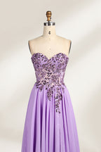 A photograph of a lavender, strapless, floor-length evening gown displayed on a dress form. The gown features intricate floral lace detailing on the bodice, transitioning into a flowing, chiffon skirt with a slight train. The dress form is positioned against a plain white background, emphasizing the gown's elegant and sophisticated design. The dress form has a wooden neck and shoulder structure with a small, round, black button at the top. The image captures the gown's luxurious texture and graceful .3
