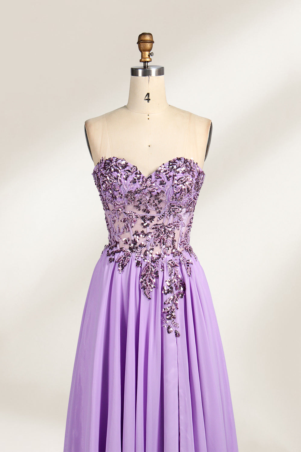 A photograph of a lavender, strapless, floor-length evening gown displayed on a dress form. The gown features intricate floral lace detailing on the bodice, transitioning into a flowing, chiffon skirt with a slight train. The dress form is positioned against a plain white background, emphasizing the gown's elegant and sophisticated design. The dress form has a wooden neck and shoulder structure with a small, round, black button at the top. The image captures the gown's luxurious texture and graceful .3