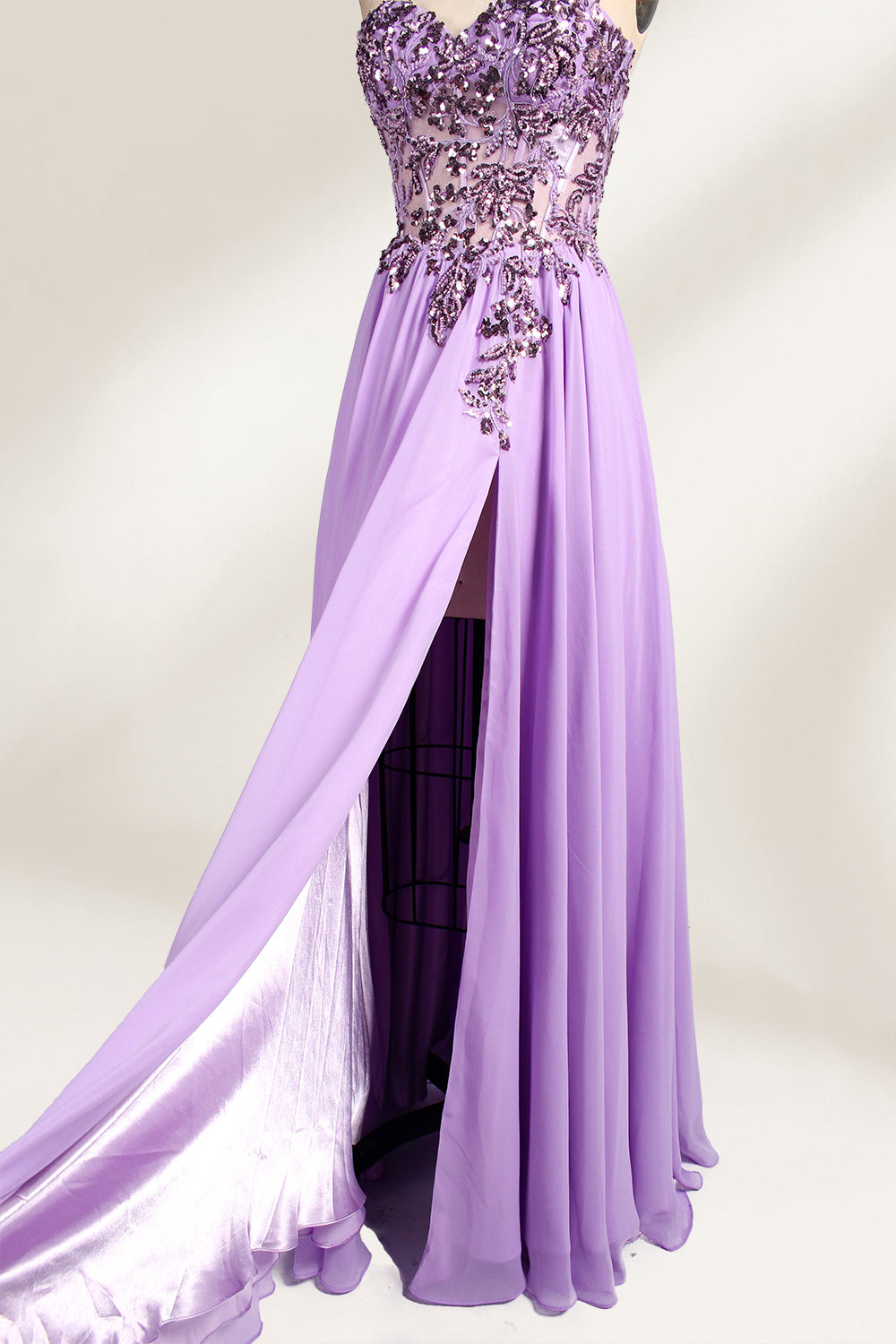 A photograph of a lavender, strapless, floor-length evening gown displayed on a dress form. The gown features intricate floral lace detailing on the bodice, transitioning into a flowing, chiffon skirt with a slight train. The dress form is positioned against a plain white background, emphasizing the gown's elegant and sophisticated design. The dress form has a wooden neck and shoulder structure with a small, round, black button at the top. The image captures the gown's luxurious texture and graceful .4
