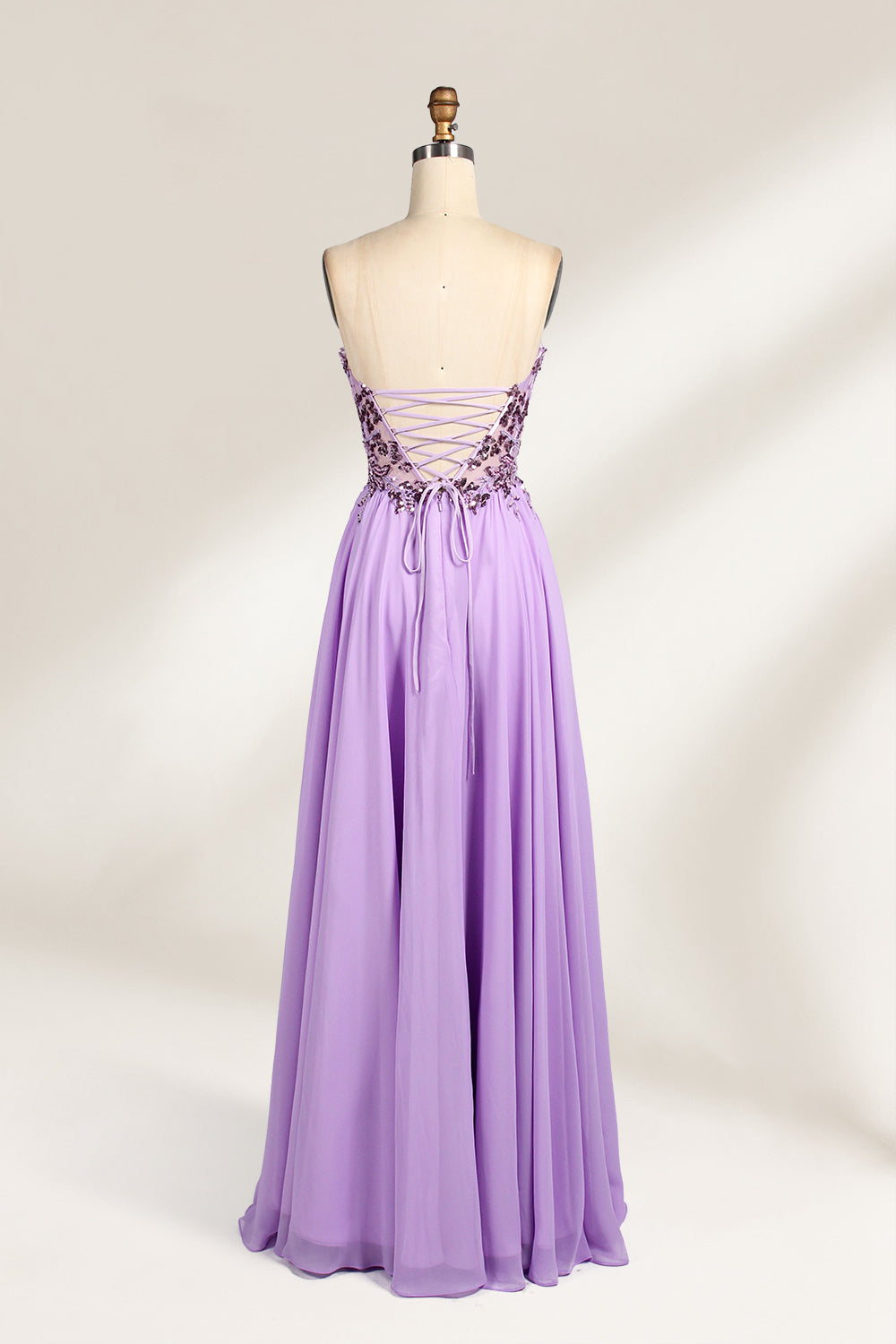 A photograph of a lavender, strapless, floor-length evening gown displayed on a dress form. The gown features intricate floral lace detailing on the bodice, transitioning into a flowing, chiffon skirt with a slight train. The dress form is positioned against a plain white background, emphasizing the gown's elegant and sophisticated design. The dress form has a wooden neck and shoulder structure with a small, round, black button at the top. The image captures the gown's luxurious texture and graceful .2
