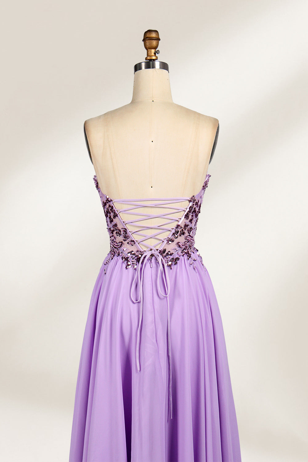 A photograph of a lavender, strapless, floor-length evening gown displayed on a dress form. The gown features intricate floral lace detailing on the bodice, transitioning into a flowing, chiffon skirt with a slight train. The dress form is positioned against a plain white background, emphasizing the gown's elegant and sophisticated design. The dress form has a wooden neck and shoulder structure with a small, round, black button at the top. The image captures the gown's luxurious texture and graceful .5