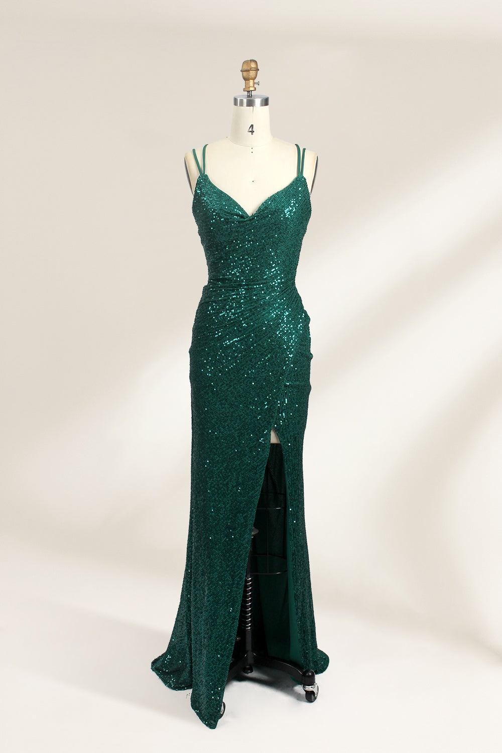 Photograph of a mannequin wearing a floor-length, deep green sequin dress with a plunging V-neckline, spaghetti straps, and a daring thigh-high slit on the left side. The dress has a fitted bodice and a flowing, slightly flared skirt. The mannequin is displayed against a plain white background with soft, diffused lighting creating gentle shadows.01