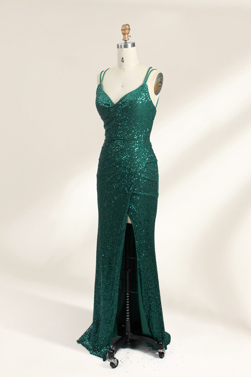 Photograph of a mannequin wearing a floor-length, deep green sequin dress with a plunging V-neckline, spaghetti straps, and a daring thigh-high slit on the left side. The dress has a fitted bodice and a flowing, slightly flared skirt. The mannequin is displayed against a plain white background with soft, diffused lighting creating gentle shadows.02