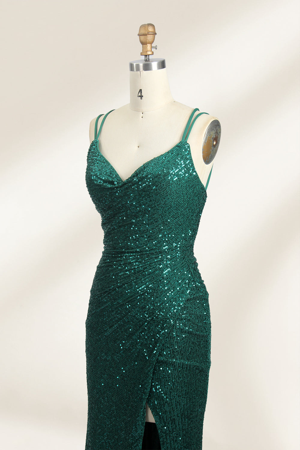 Photograph of a mannequin wearing a floor-length, deep green sequin dress with a plunging V-neckline, spaghetti straps, and a daring thigh-high slit on the left side. The dress has a fitted bodice and a flowing, slightly flared skirt. The mannequin is displayed against a plain white background with soft, diffused lighting creating gentle shadows.03