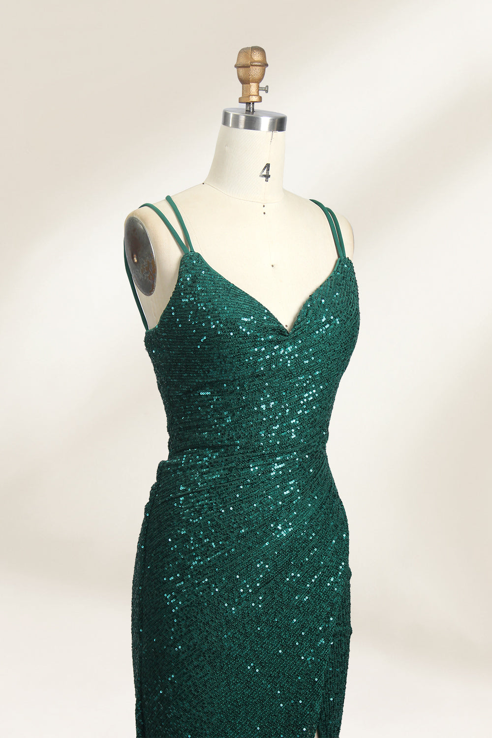 Photograph of a mannequin wearing a floor-length, deep green sequin dress with a plunging V-neckline, spaghetti straps, and a daring thigh-high slit on the left side. The dress has a fitted bodice and a flowing, slightly flared skirt. The mannequin is displayed against a plain white background with soft, diffused lighting creating gentle shadows.04