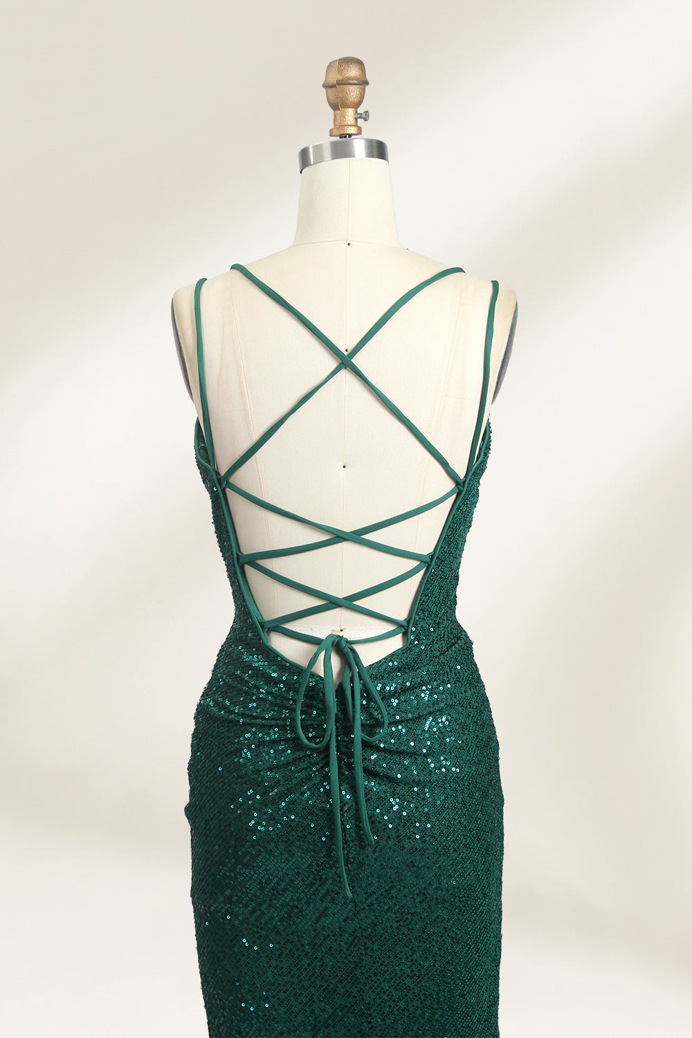 Photograph of a mannequin wearing a floor-length, deep green sequin dress with a plunging V-neckline, spaghetti straps, and a daring thigh-high slit on the left side. The dress has a fitted bodice and a flowing, slightly flared skirt. The mannequin is displayed against a plain white background with soft, diffused lighting creating gentle shadows.06