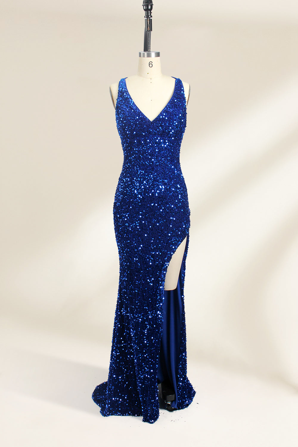 Photograph of a mannequin wearing a floor-length, deep V-neck, navy blue sequin dress with a high slit on the right side. The dress has a fitted bodice and a flared skirt. The background is a plain, light beige. The mannequin stands on a white pedestal, and the dress sparkles under soft, diffused lighting.01