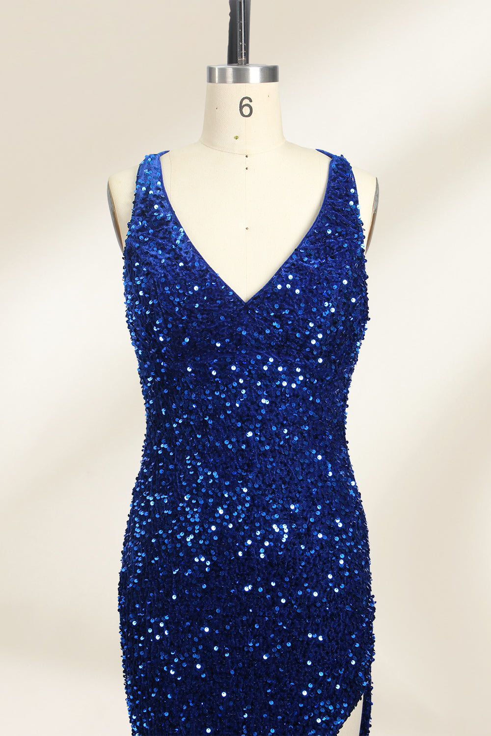 Photograph of a mannequin wearing a floor-length, deep V-neck, navy blue sequin dress with a high slit on the right side. The dress has a fitted bodice and a flared skirt. The background is a plain, light beige. The mannequin stands on a white pedestal, and the dress sparkles under soft, diffused lighting.03