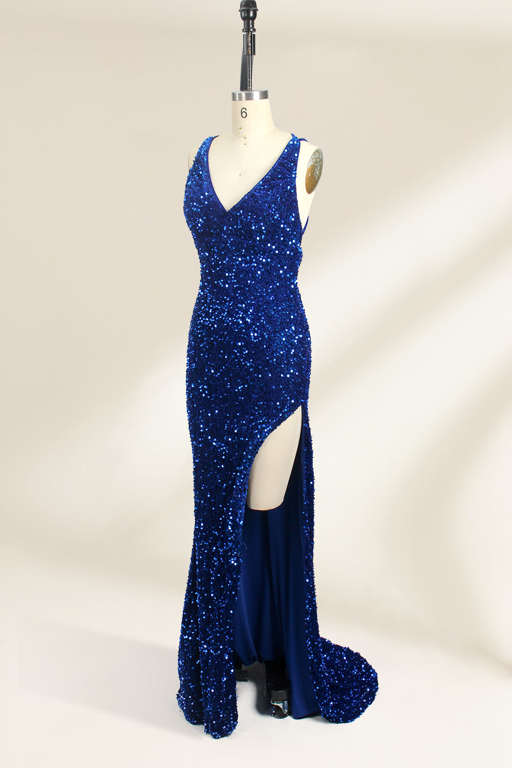 Photograph of a mannequin wearing a floor-length, deep V-neck, navy blue sequin dress with a high slit on the right side. The dress has a fitted bodice and a flared skirt. The background is a plain, light beige. The mannequin stands on a white pedestal, and the dress sparkles under soft, diffused lighting.04