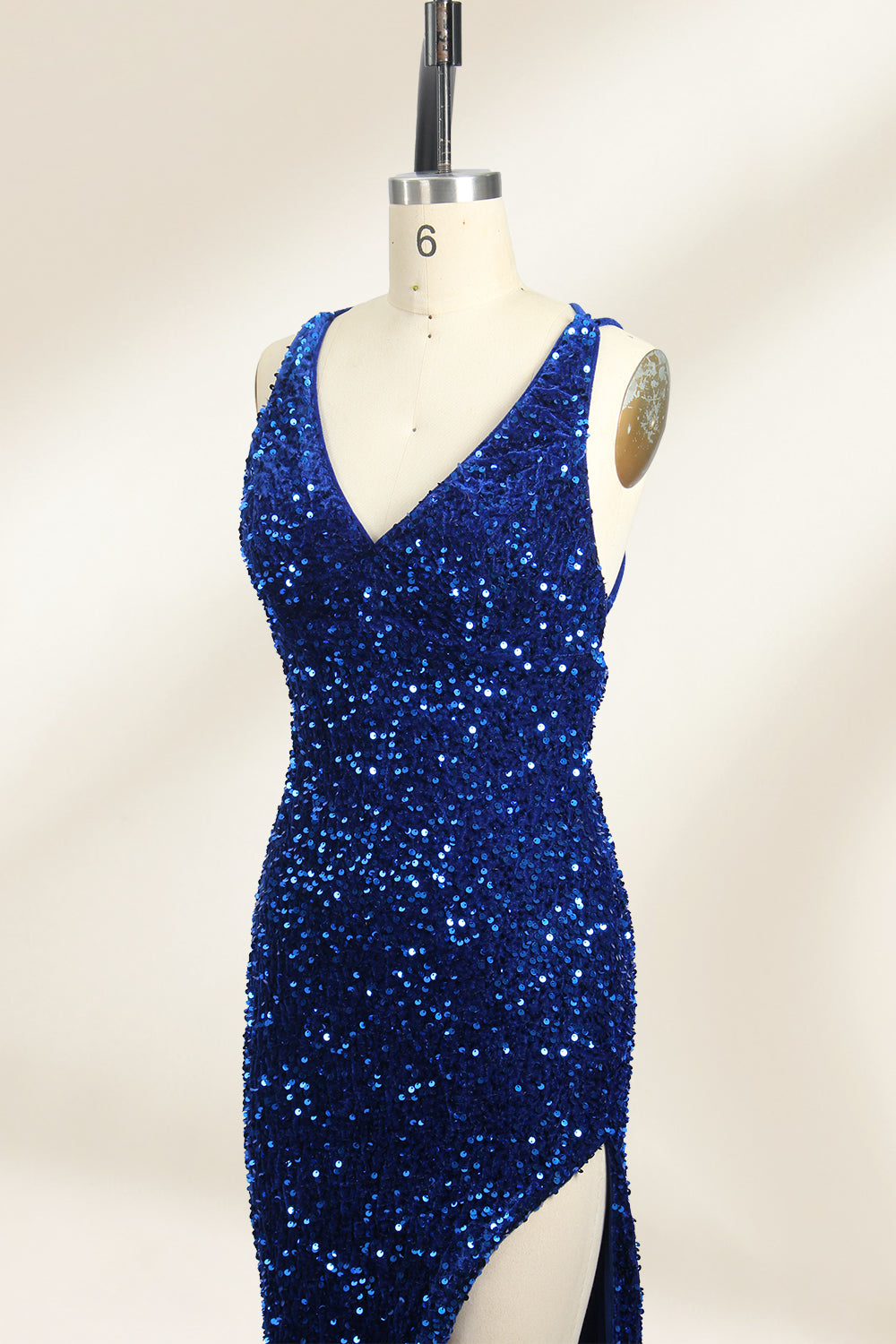 Photograph of a mannequin wearing a floor-length, deep V-neck, navy blue sequin dress with a high slit on the right side. The dress has a fitted bodice and a flared skirt. The background is a plain, light beige. The mannequin stands on a white pedestal, and the dress sparkles under soft, diffused lighting.06