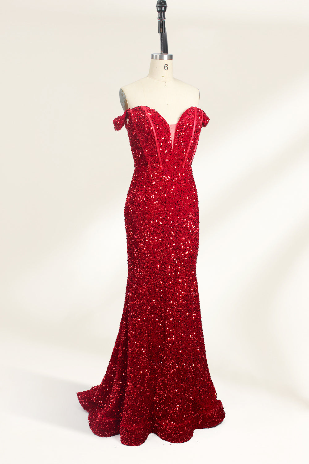 A photograph of a mannequin showcasing an off-the-shoulder, floor-length red sequin gown with a plunging V-neckline. The dress has a fitted bodice and a flared skirt, creating a glamorous, elegant appearance. The mannequin stands against a plain white background, emphasizing the dress's vibrant color and intricate details.03