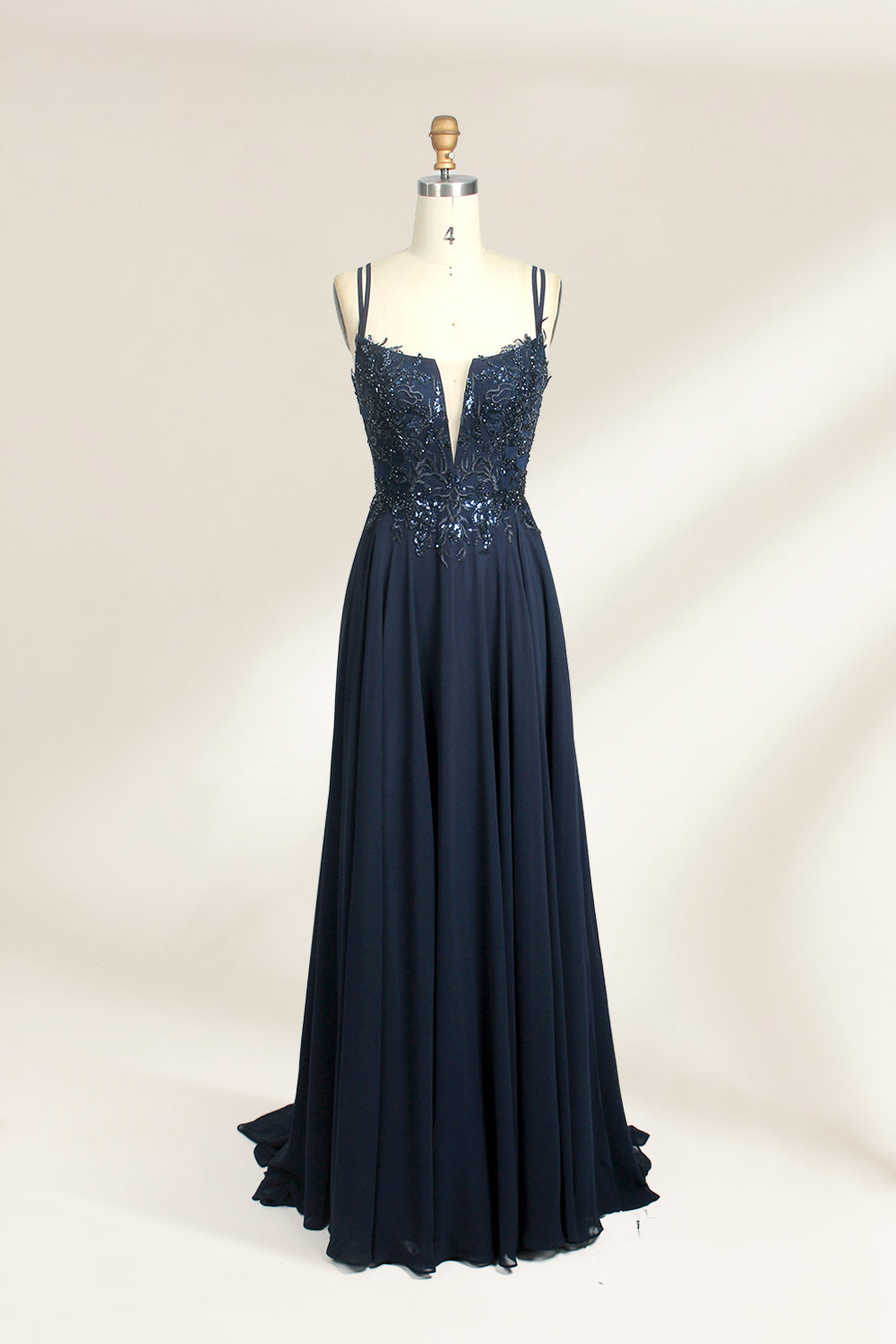 Photograph of a mannequin showcasing a floor-length, dark navy blue evening gown. The gown features a plunging V-neckline adorned with intricate beadwork and sequins. The fabric is a flowing chiffon, creating soft, elegant draping. The mannequin stands against a plain, light beige background, emphasizing the gown's luxurious texture and design. The mannequin's head is replaced with a wooden headpiece, and the dress is fitted to highlight the elegant, sophisticated style.01