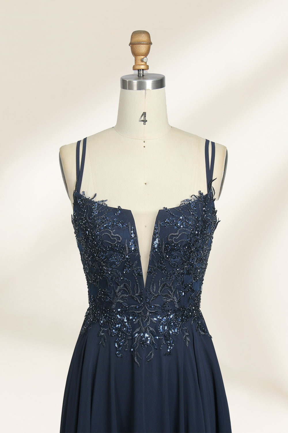 Photograph of a mannequin showcasing a floor-length, dark navy blue evening gown. The gown features a plunging V-neckline adorned with intricate beadwork and sequins. The fabric is a flowing chiffon, creating soft, elegant draping. The mannequin stands against a plain, light beige background, emphasizing the gown's luxurious texture and design. The mannequin's head is replaced with a wooden headpiece, and the dress is fitted to highlight the elegant, sophisticated style.04