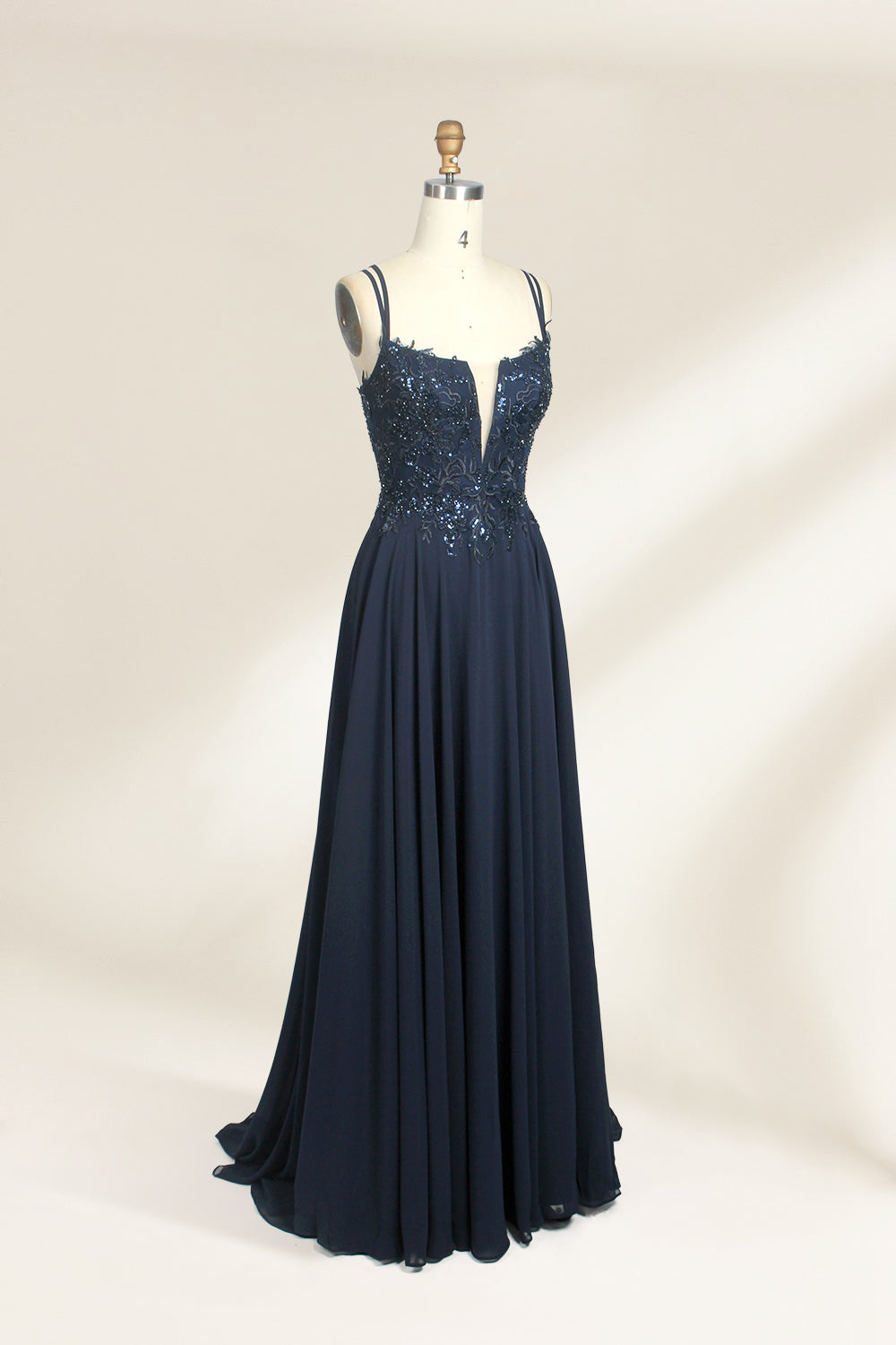 Photograph of a mannequin showcasing a floor-length, dark navy blue evening gown. The gown features a plunging V-neckline adorned with intricate beadwork and sequins. The fabric is a flowing chiffon, creating soft, elegant draping. The mannequin stands against a plain, light beige background, emphasizing the gown's luxurious texture and design. The mannequin's head is replaced with a wooden headpiece, and the dress is fitted to highlight the elegant, sophisticated style.05