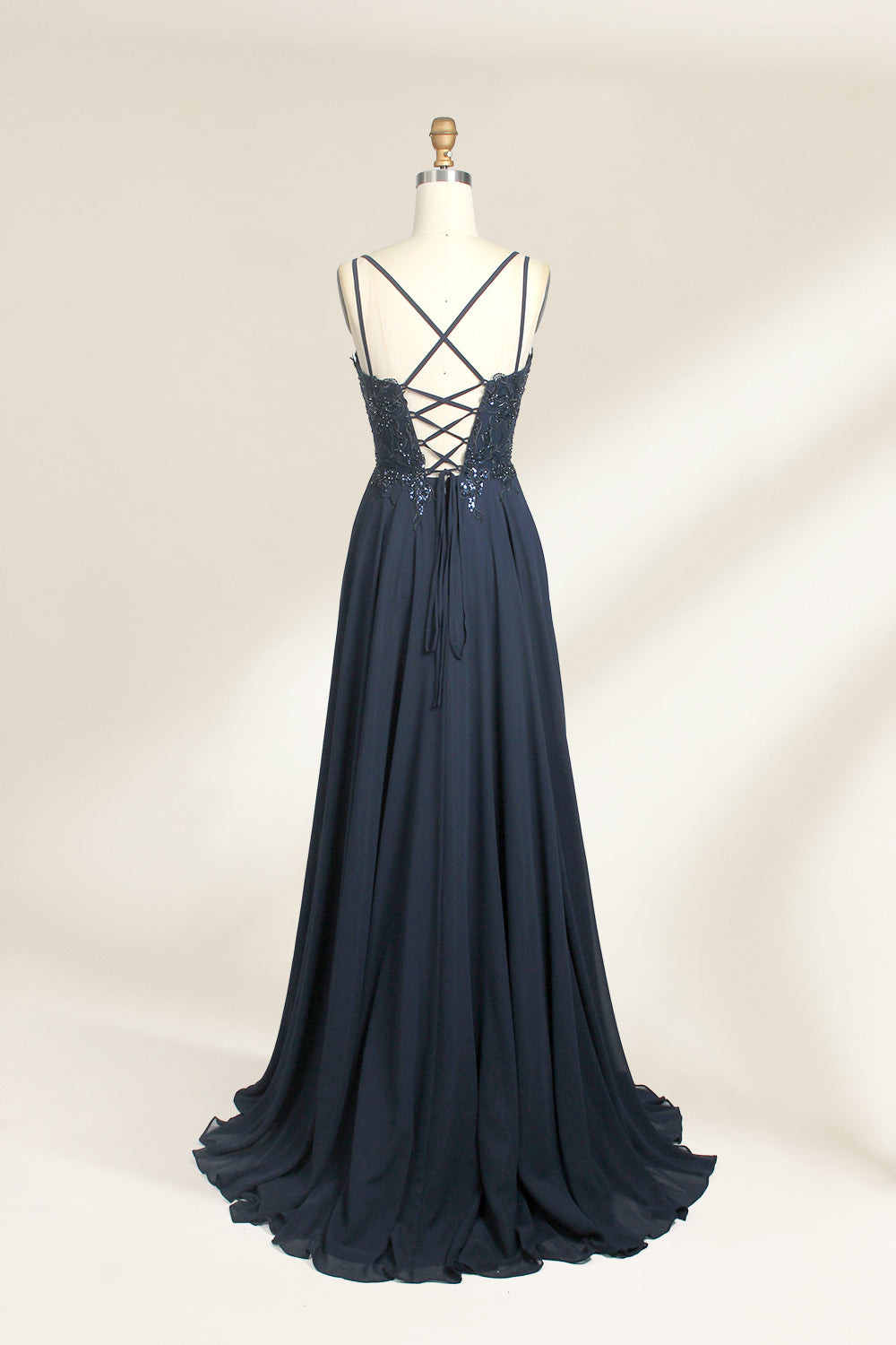 Photograph of a mannequin showcasing a floor-length, dark navy blue evening gown. The gown features a plunging V-neckline adorned with intricate beadwork and sequins. The fabric is a flowing chiffon, creating soft, elegant draping. The mannequin stands against a plain, light beige background, emphasizing the gown's luxurious texture and design. The mannequin's head is replaced with a wooden headpiece, and the dress is fitted to highlight the elegant, sophisticated style.02