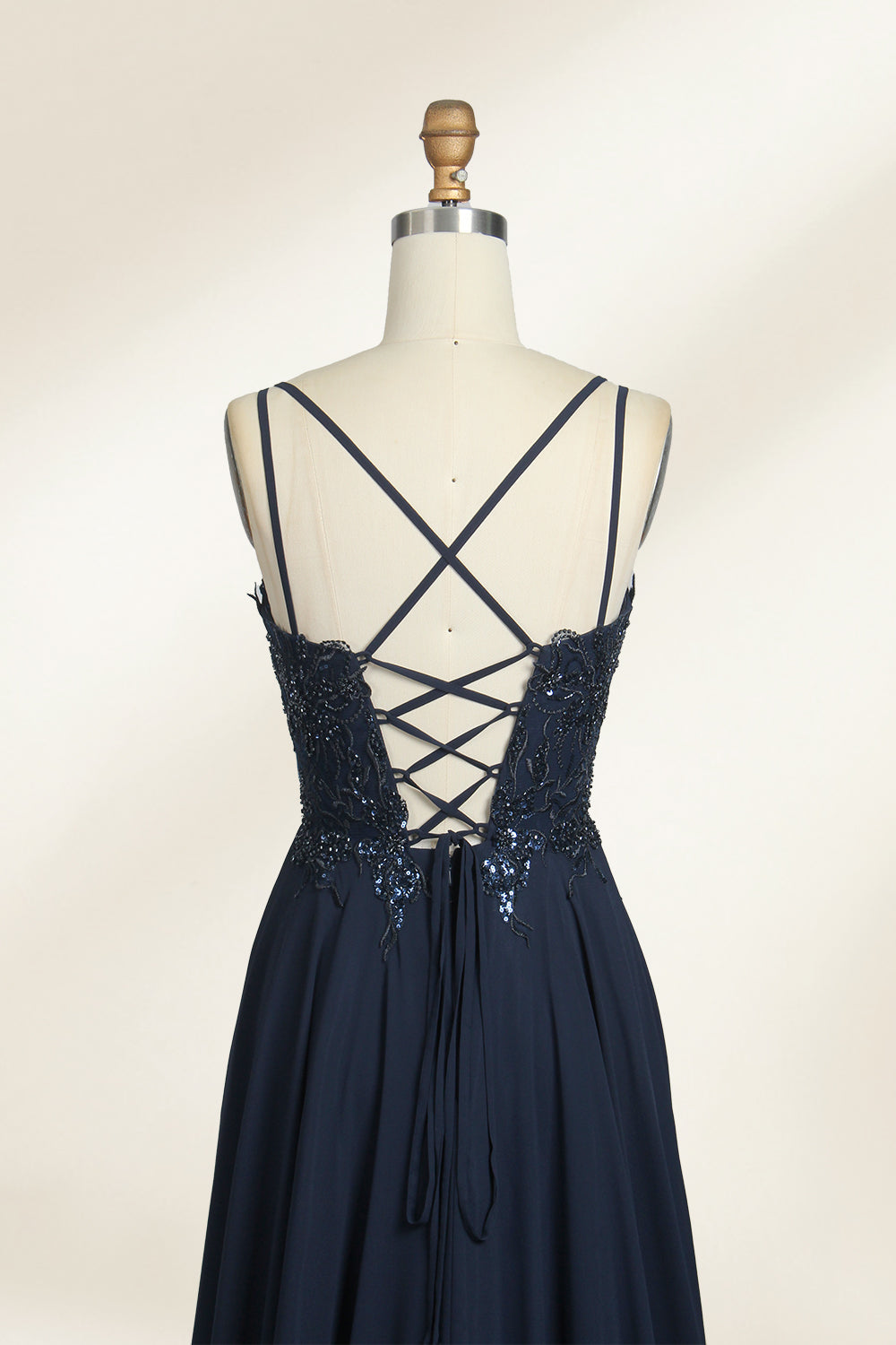 Photograph of a mannequin showcasing a floor-length, dark navy blue evening gown. The gown features a plunging V-neckline adorned with intricate beadwork and sequins. The fabric is a flowing chiffon, creating soft, elegant draping. The mannequin stands against a plain, light beige background, emphasizing the gown's luxurious texture and design. The mannequin's head is replaced with a wooden headpiece, and the dress is fitted to highlight the elegant, sophisticated style.07