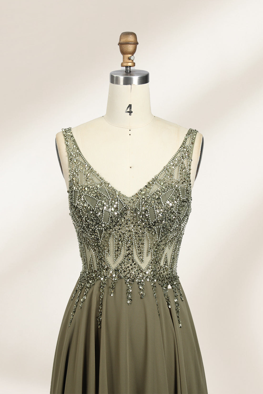 Photograph of an elegant, sleeveless, floor-length gown displayed on a dress form. The gown is a rich olive green with a fitted bodice adorned with intricate, sparkling embellishments. The neckline is a deep V, and the waistline is subtly cinched. The dress flows into a soft, gathered skirt. The background is a plain, light beige, enhancing the gown's luxurious texture and shimmering details.03