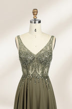 Photograph of an elegant, sleeveless, floor-length gown displayed on a dress form. The gown is a rich olive green with a fitted bodice adorned with intricate, sparkling embellishments. The neckline is a deep V, and the waistline is subtly cinched. The dress flows into a soft, gathered skirt. The background is a plain, light beige, enhancing the gown's luxurious texture and shimmering details.03