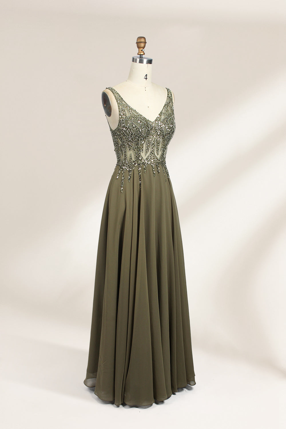 Photograph of an elegant, sleeveless, floor-length gown displayed on a dress form. The gown is a rich olive green with a fitted bodice adorned with intricate, sparkling embellishments. The neckline is a deep V, and the waistline is subtly cinched. The dress flows into a soft, gathered skirt. The background is a plain, light beige, enhancing the gown's luxurious texture and shimmering details.04