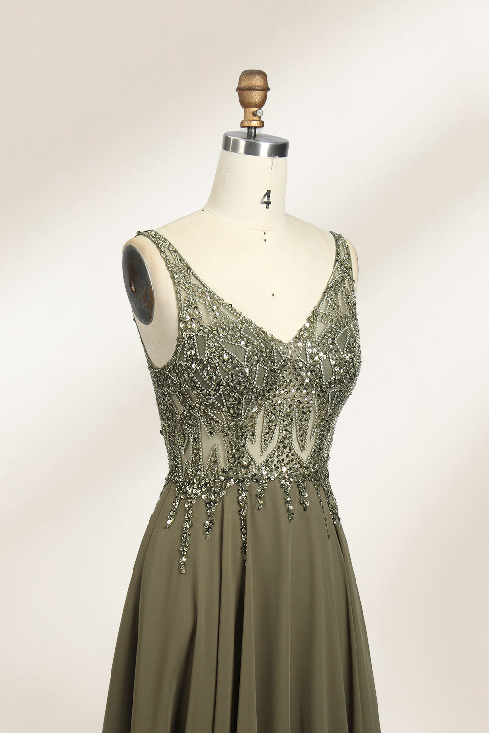 Photograph of an elegant, sleeveless, floor-length gown displayed on a dress form. The gown is a rich olive green with a fitted bodice adorned with intricate, sparkling embellishments. The neckline is a deep V, and the waistline is subtly cinched. The dress flows into a soft, gathered skirt. The background is a plain, light beige, enhancing the gown's luxurious texture and shimmering details.05