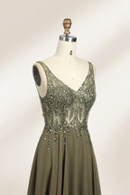 Photograph of an elegant, sleeveless, floor-length gown displayed on a dress form. The gown is a rich olive green with a fitted bodice adorned with intricate, sparkling embellishments. The neckline is a deep V, and the waistline is subtly cinched. The dress flows into a soft, gathered skirt. The background is a plain, light beige, enhancing the gown's luxurious texture and shimmering details.05
