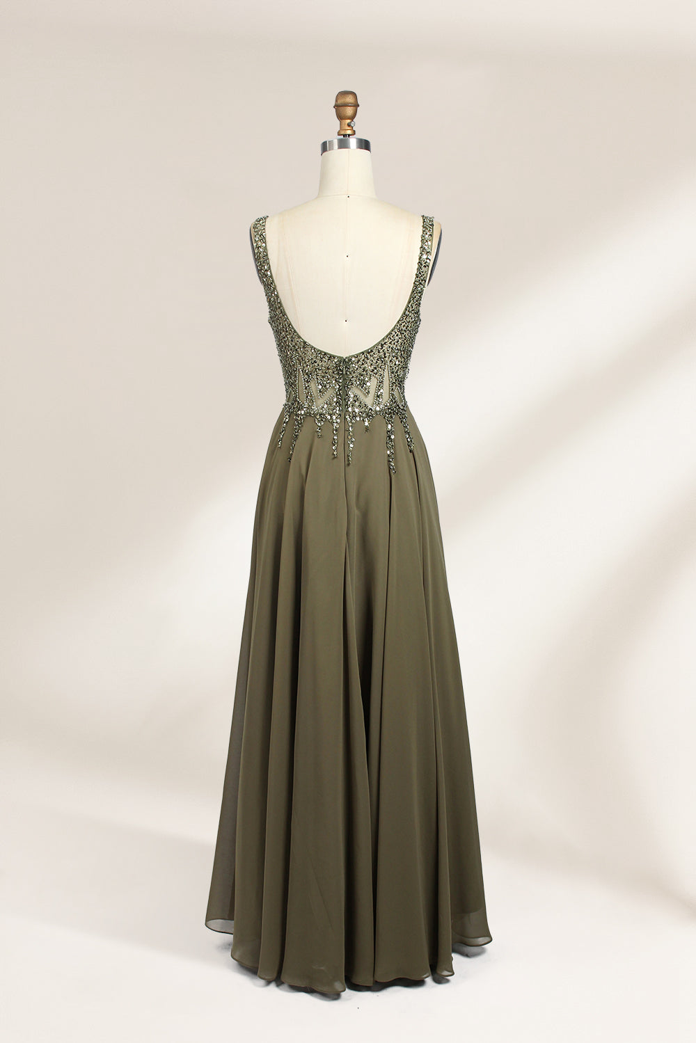 Photograph of an elegant, sleeveless, floor-length gown displayed on a dress form. The gown is a rich olive green with a fitted bodice adorned with intricate, sparkling embellishments. The neckline is a deep V, and the waistline is subtly cinched. The dress flows into a soft, gathered skirt. The background is a plain, light beige, enhancing the gown's luxurious texture and shimmering details.02