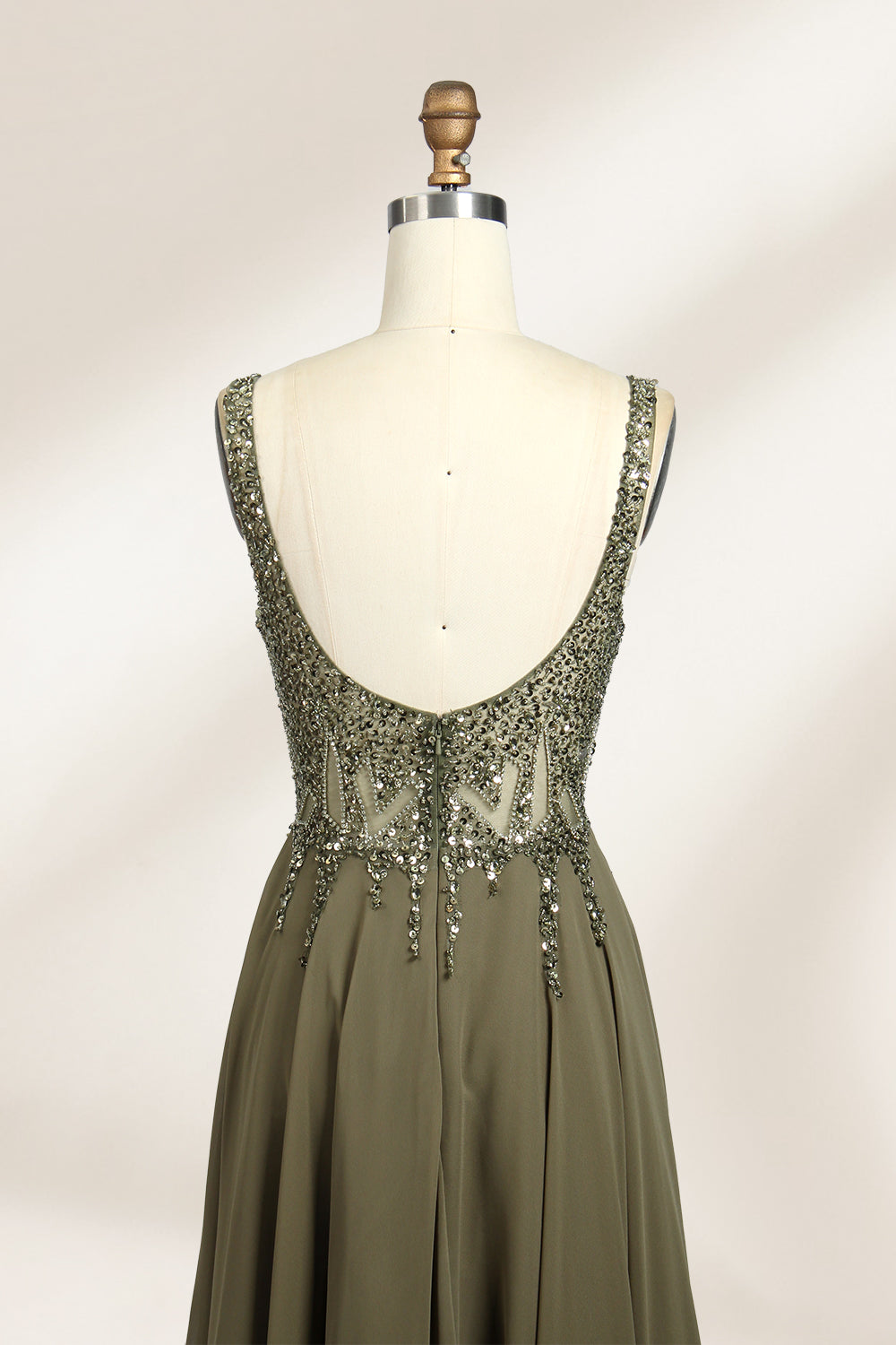 Photograph of an elegant, sleeveless, floor-length gown displayed on a dress form. The gown is a rich olive green with a fitted bodice adorned with intricate, sparkling embellishments. The neckline is a deep V, and the waistline is subtly cinched. The dress flows into a soft, gathered skirt. The background is a plain, light beige, enhancing the gown's luxurious texture and shimmering details.06