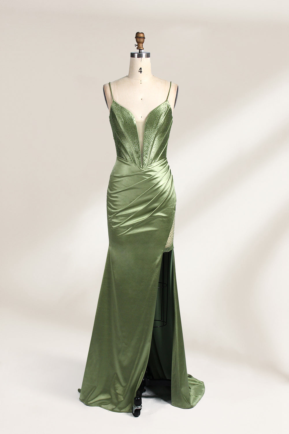 A mannequin showcases an olive - green satin gown. It has thin straps, a deep V - neck, ruched bodice, and a high side slit with sheer details. The elegant design, blending satin and intricate ruching, suits formal events like galas or proms, radiating sophistication.1