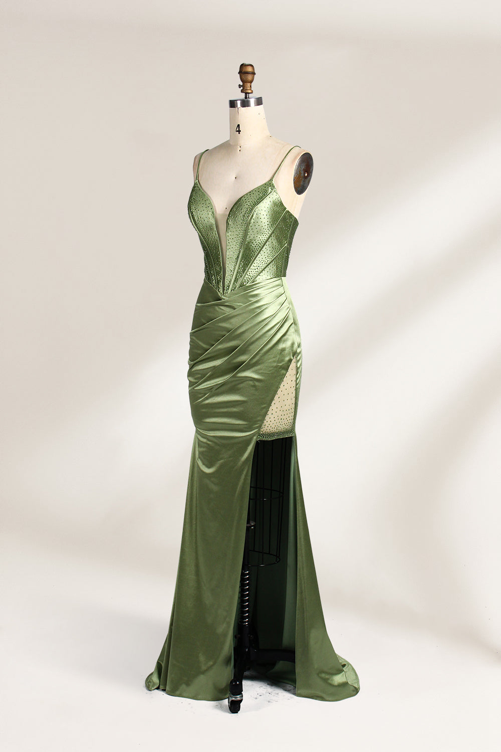 A mannequin showcases an olive - green satin gown. It has thin straps, a deep V - neck, ruched bodice, and a high side slit with sheer details. The elegant design, blending satin and intricate ruching, suits formal events like galas or proms, radiating sophistication.4