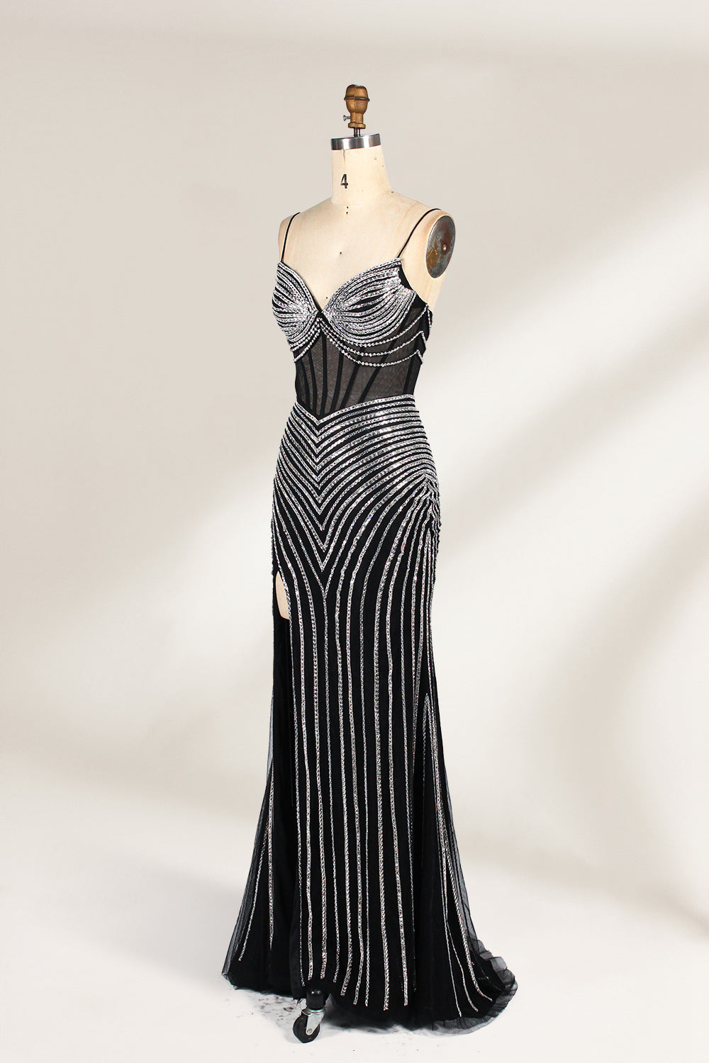 A mannequin showcases a black gown with silver beading. It has thin straps, a sheer bodice, and a high side slit. The geometric beading pattern adds glamour, making it perfect for formal events like galas or proms, radiating sophistication.4