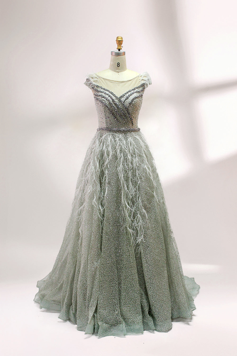 Displayed on a mannequin is a light - green evening gown. It features a sheer neckline with feather and bead embellishments, and a voluminous, glittering skirt. The elegant design creates a dreamy and luxurious look, perfect for formal events.1