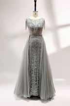 Displayed on a mannequin is a silver - gray evening gown. It features a sheer neckline with fringe details, intricate lace embroidery on the bodice, and a flowing tulle overlay. The elegant design creates a sophisticated and glamorous look, perfect for formal events.1