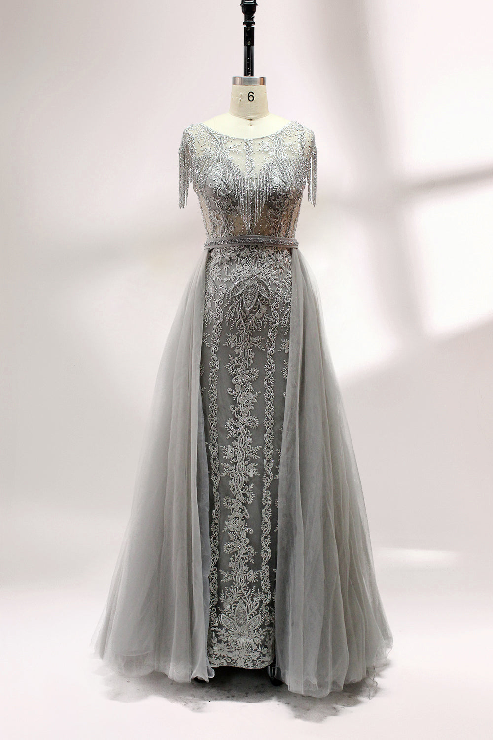 Displayed on a mannequin is a silver - gray evening gown. It features a sheer neckline with fringe details, intricate lace embroidery on the bodice, and a flowing tulle overlay. The elegant design creates a sophisticated and glamorous look, perfect for formal events.1