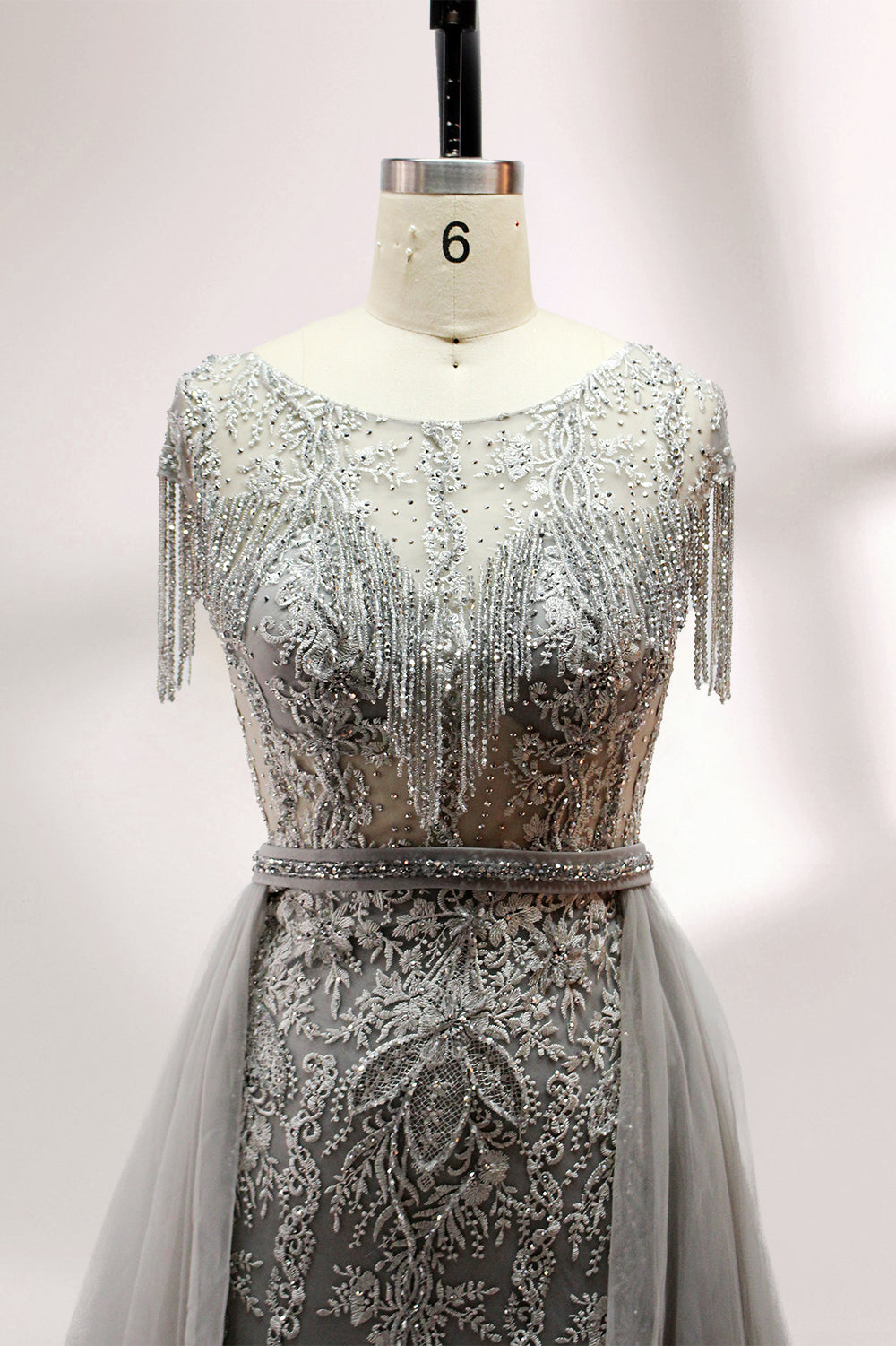 Displayed on a mannequin is a silver - gray evening gown. It features a sheer neckline with fringe details, intricate lace embroidery on the bodice, and a flowing tulle overlay. The elegant design creates a sophisticated and glamorous look, perfect for formal events.4