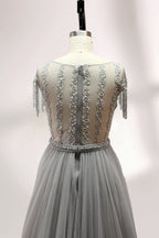Displayed on a mannequin is a silver - gray evening gown. It features a sheer neckline with fringe details, intricate lace embroidery on the bodice, and a flowing tulle overlay. The elegant design creates a sophisticated and glamorous look, perfect for formal events.5