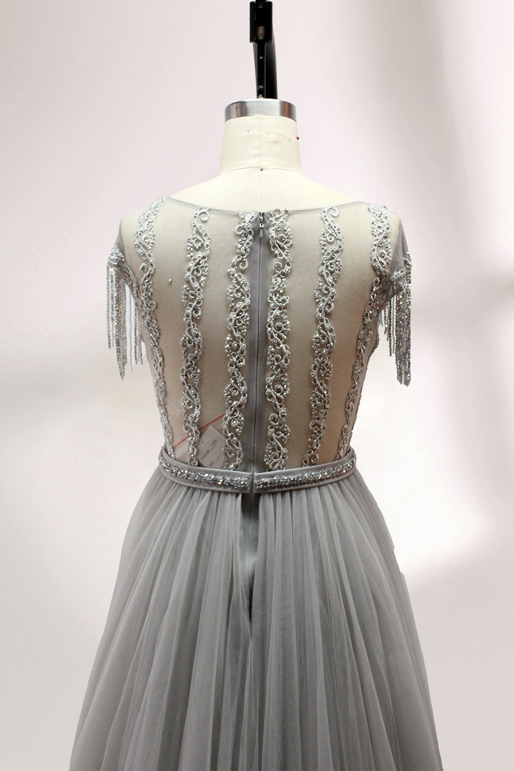 Displayed on a mannequin is a silver - gray evening gown. It features a sheer neckline with fringe details, intricate lace embroidery on the bodice, and a flowing tulle overlay. The elegant design creates a sophisticated and glamorous look, perfect for formal events.5