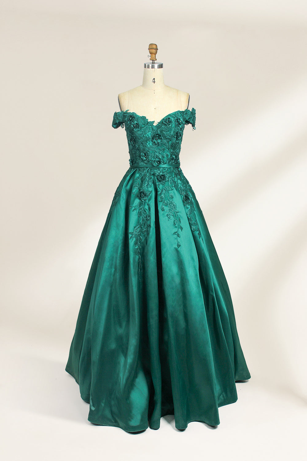 This is a photograph of a stunning, off-the-shoulder, floor-length evening gown displayed on a dress form. The gown is a rich, emerald green satin with intricate black floral lace embroidery on the bodice and hem. The dress features a sweetheart neckline, accentuating the bust, and a fitted waist that flares into a full, flowing skirt. The dress form is beige, with a wooden head and neck, and a metal neck ring. The background is a plain, light cream color, ensuring the dress remains the focal point.01