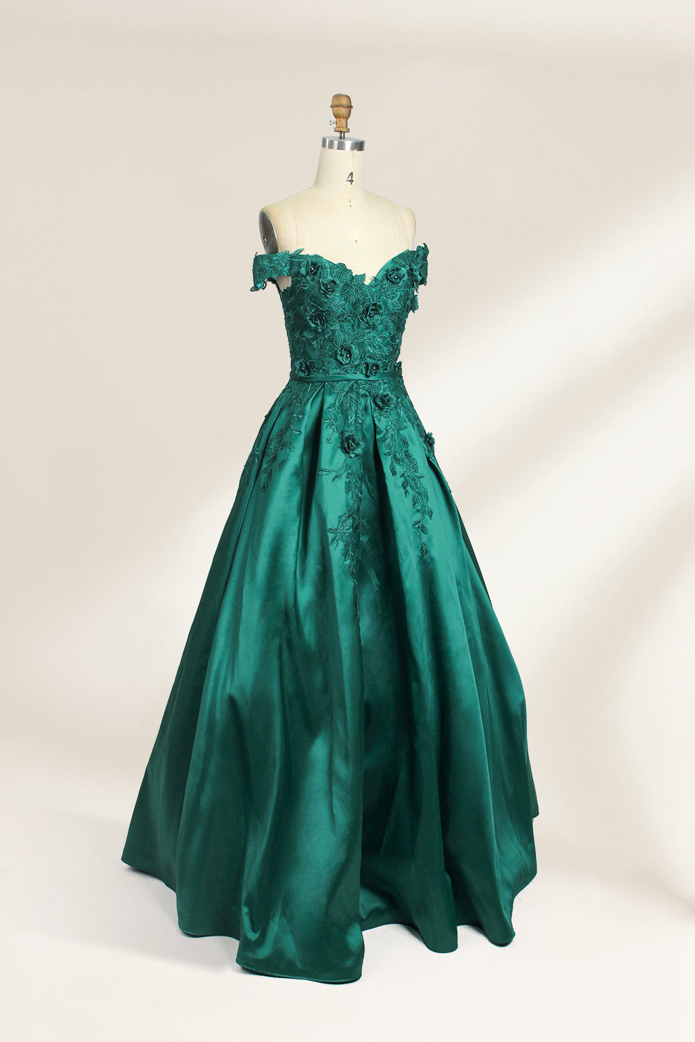 This is a photograph of a stunning, off-the-shoulder, floor-length evening gown displayed on a dress form. The gown is a rich, emerald green satin with intricate black floral lace embroidery on the bodice and hem. The dress features a sweetheart neckline, accentuating the bust, and a fitted waist that flares into a full, flowing skirt. The dress form is beige, with a wooden head and neck, and a metal neck ring. The background is a plain, light cream color, ensuring the dress remains the focal point.03