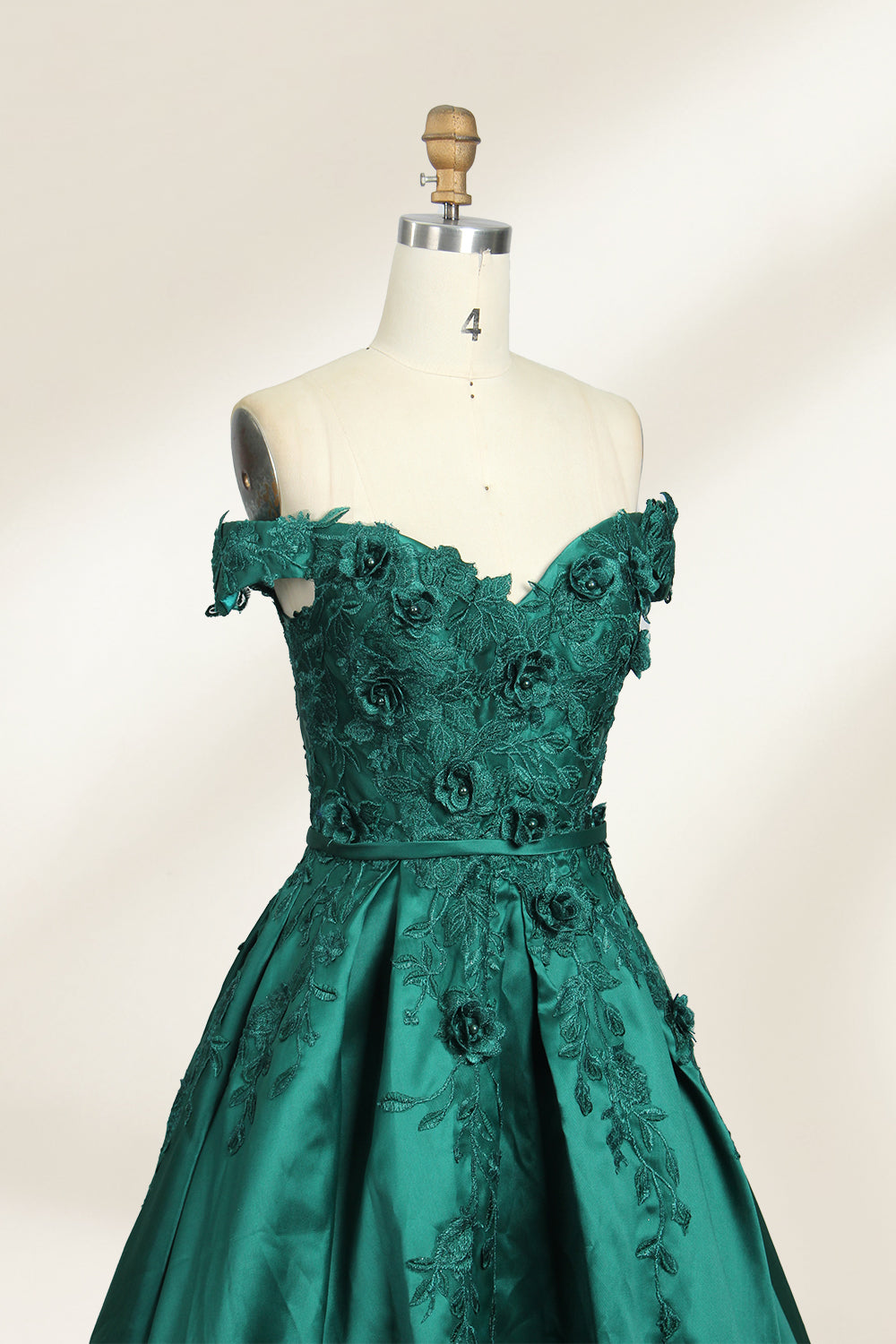 This is a photograph of a stunning, off-the-shoulder, floor-length evening gown displayed on a dress form. The gown is a rich, emerald green satin with intricate black floral lace embroidery on the bodice and hem. The dress features a sweetheart neckline, accentuating the bust, and a fitted waist that flares into a full, flowing skirt. The dress form is beige, with a wooden head and neck, and a metal neck ring. The background is a plain, light cream color, ensuring the dress remains the focal point.04