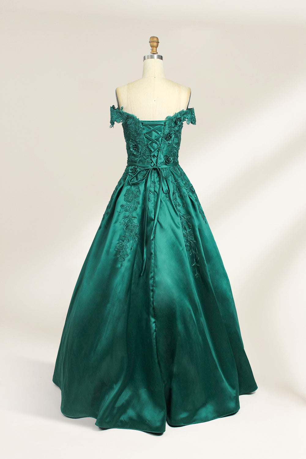 This is a photograph of a stunning, off-the-shoulder, floor-length evening gown displayed on a dress form. The gown is a rich, emerald green satin with intricate black floral lace embroidery on the bodice and hem. The dress features a sweetheart neckline, accentuating the bust, and a fitted waist that flares into a full, flowing skirt. The dress form is beige, with a wooden head and neck, and a metal neck ring. The background is a plain, light cream color, ensuring the dress remains the focal point.02