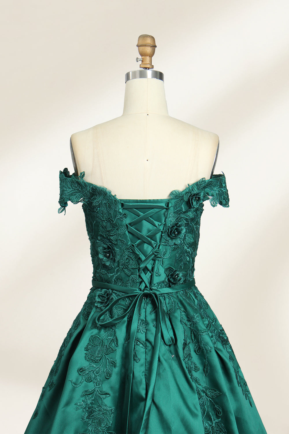 This is a photograph of a stunning, off-the-shoulder, floor-length evening gown displayed on a dress form. The gown is a rich, emerald green satin with intricate black floral lace embroidery on the bodice and hem. The dress features a sweetheart neckline, accentuating the bust, and a fitted waist that flares into a full, flowing skirt. The dress form is beige, with a wooden head and neck, and a metal neck ring. The background is a plain, light cream color, ensuring the dress remains the focal point.05
