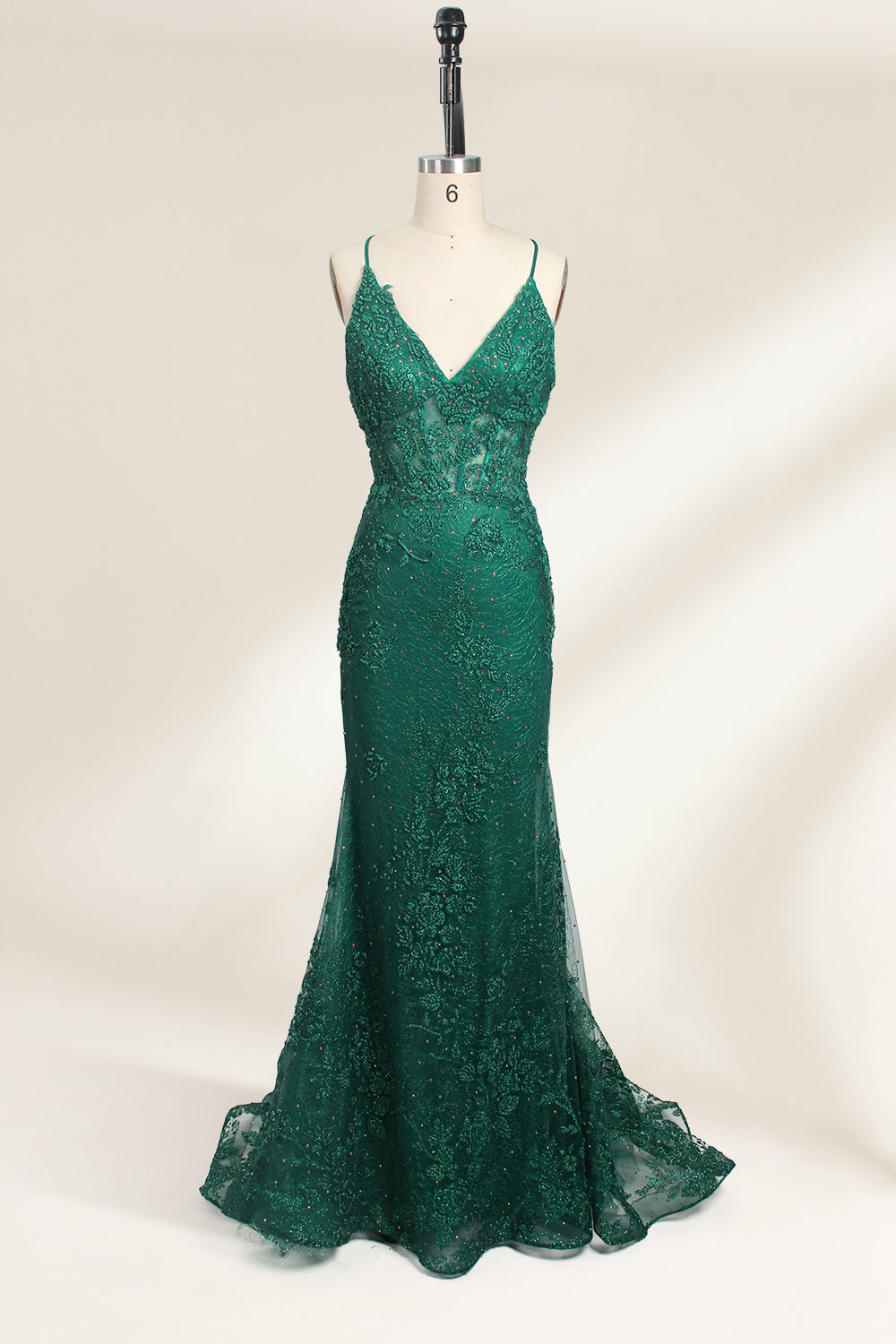 Photograph of a dark green, floor-length evening gown displayed on a white dress form. The gown features a deep V-neckline with thin spaghetti straps, a fitted bodice adorned with intricate lace detailing, and a flowing, slightly flared skirt. The dress showcases a rich, textured fabric with a shimmering effect, accentuating the luxurious and elegant style. The background is a plain, off-white color, ensuring the dress remains the focal point.01