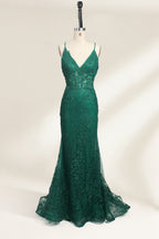Photograph of a dark green, floor-length evening gown displayed on a white dress form. The gown features a deep V-neckline with thin spaghetti straps, a fitted bodice adorned with intricate lace detailing, and a flowing, slightly flared skirt. The dress showcases a rich, textured fabric with a shimmering effect, accentuating the luxurious and elegant style. The background is a plain, off-white color, ensuring the dress remains the focal point.01
